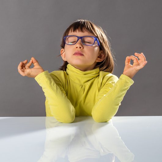 little yoga child with eyeglasses breathing, practicing yoga and closing eyes with inner balance hand gesture at the table for zen and health at school, grey background