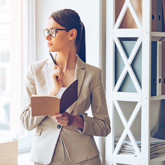 I need to make some changes! Pensive young beautiful businesswoman in glasses holding notebook and looking through window while standing at her working place