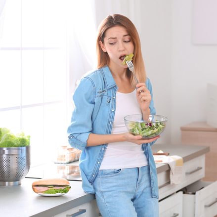 Emotional young woman eating salad instead of sandwich in kitchen. Healthy diet