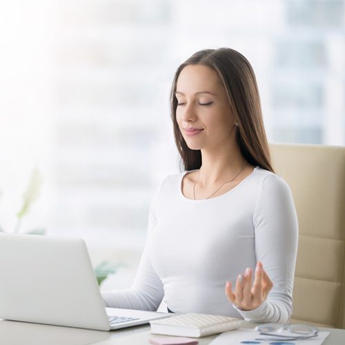 Young woman near the laptop, practicing meditation at the office desk, in front of laptop, online yoga classes, taking a break time for a minute, healing from paperwork and laptop radiation